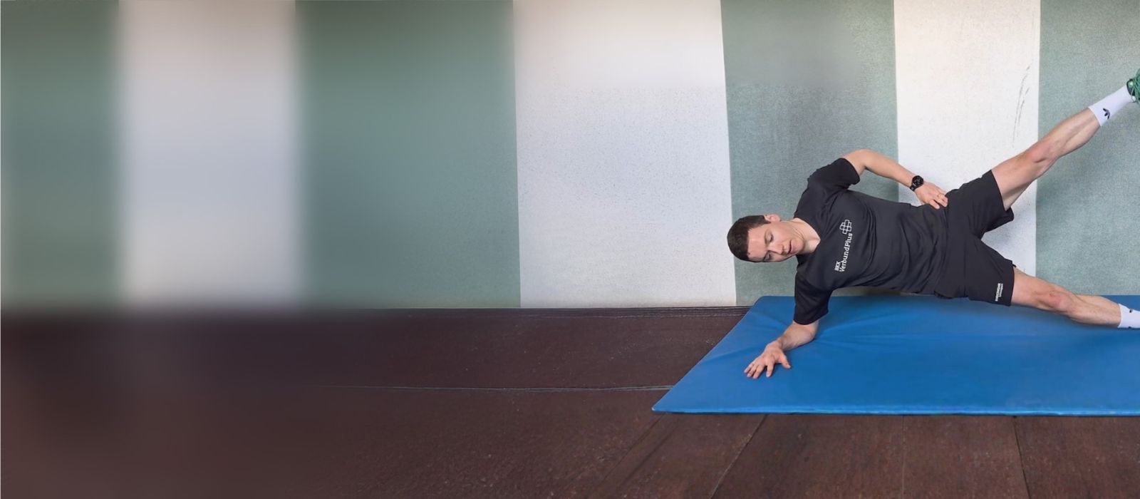 Ein Mann führt auf einer blauen Fitnessmatte in einem Innenraum eine seitliche Plank-Variation aus. Er stützt sich auf einen Unterarm, der Körper ist seitlich gestreckt, ein Bein ist angehoben. Der Raum hat einen dunklen Holzboden und eine hellgrün-weiß gestreifte Wand.