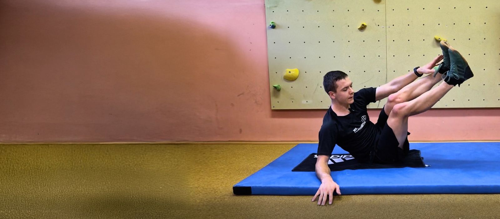 Justus Strelow auf einer Trainingsmatte bei einer anspruchsvollen Rumpfübung mit angehobenen Beinen in einer Sporthalle