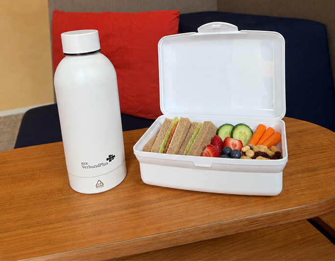 Gesunde Vesperbox für Kinder: Neben einer weißen Trinkflasche steht eine geöffnete Lunchbox mit belegten Vollkornbroten, Gurkenscheiben, Erdbeeren, Heidelbeeren, Möhren, Nüssen und Trockenfrüchten auf einem Holztisch.